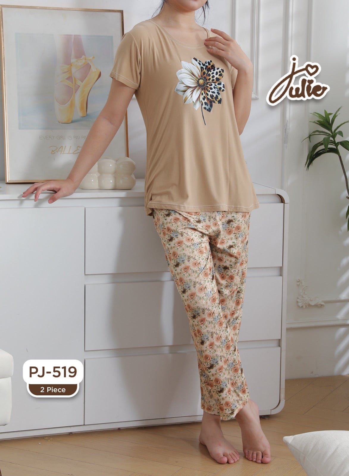 A woman modeling a two-piece pajama set consisting of a beige short-sleeve top with a white and black floral print, and matching floral pajama pants. The setting is a bright bedroom with a white dresser, framed art, and a plant.