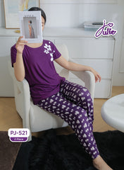 A woman seated in a white armchair wearing a purple two-piece pajama set with a floral print. She holds a magazine in front of her face.