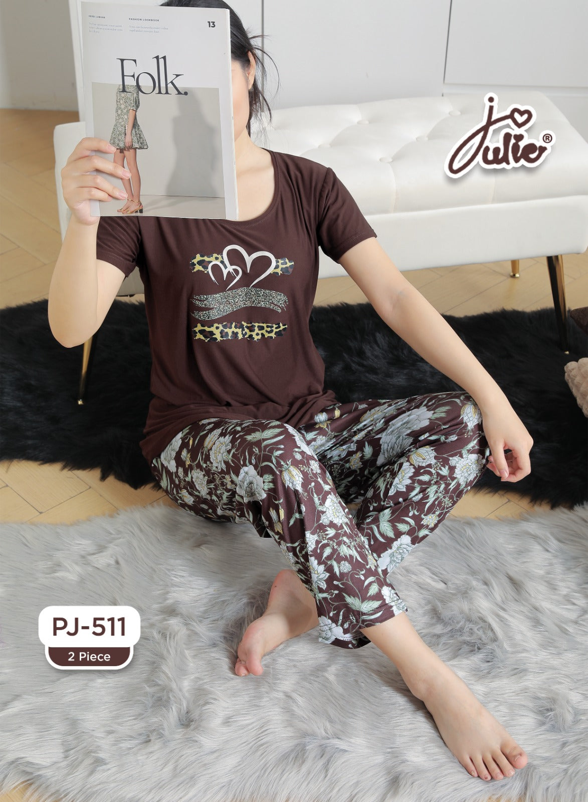 A woman sits cross-legged on a gray faux-fur rug in a living room setting, wearing a two-piece pajama set consisting of a dark brown short-sleeve top with a heart graphic and matching floral-print pants. She is holding a magazine, and a white tufted sofa is visible in the background.