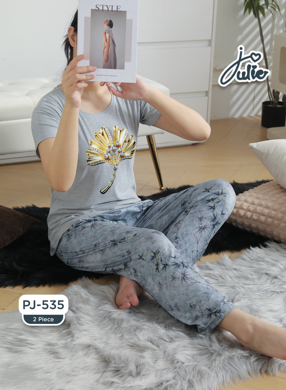 A woman sitting on a white faux fur rug in a bedroom setting, wearing a light gray t-shirt with a gold sun graphic and matching light blue patterned pajama pants. She is holding a magazine in front of her face.
