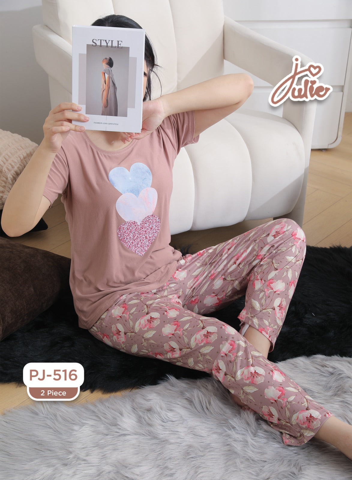 A woman wearing a two-piece pajama set sits on a white armchair in a living room setting. The pajama set consists of a dusty pink short-sleeve top with a heart graphic and matching floral-printed pants.
