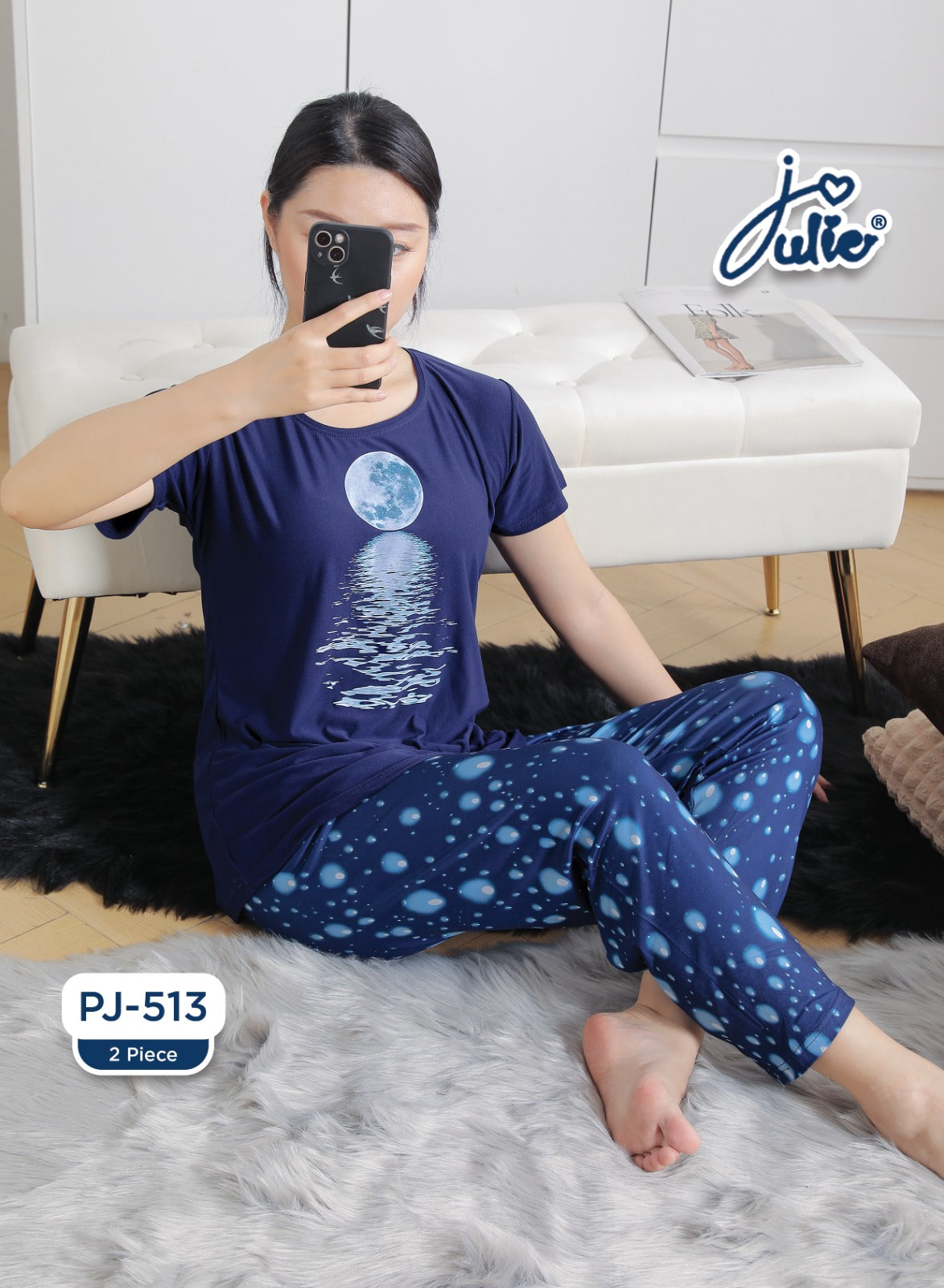 A woman wearing a navy blue two-piece pajama set with a moon graphic on the t-shirt and blue polka-dot patterned pants, sitting on a white faux-fur rug in front of a white tufted bench.