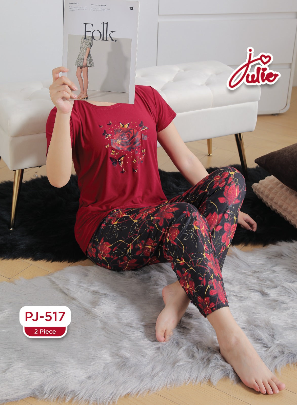A woman sits barefoot on a gray rug in a bright room, wearing a red short-sleeve top with a floral graphic and matching black floral pajama pants. She holds a magazine in front of her face.